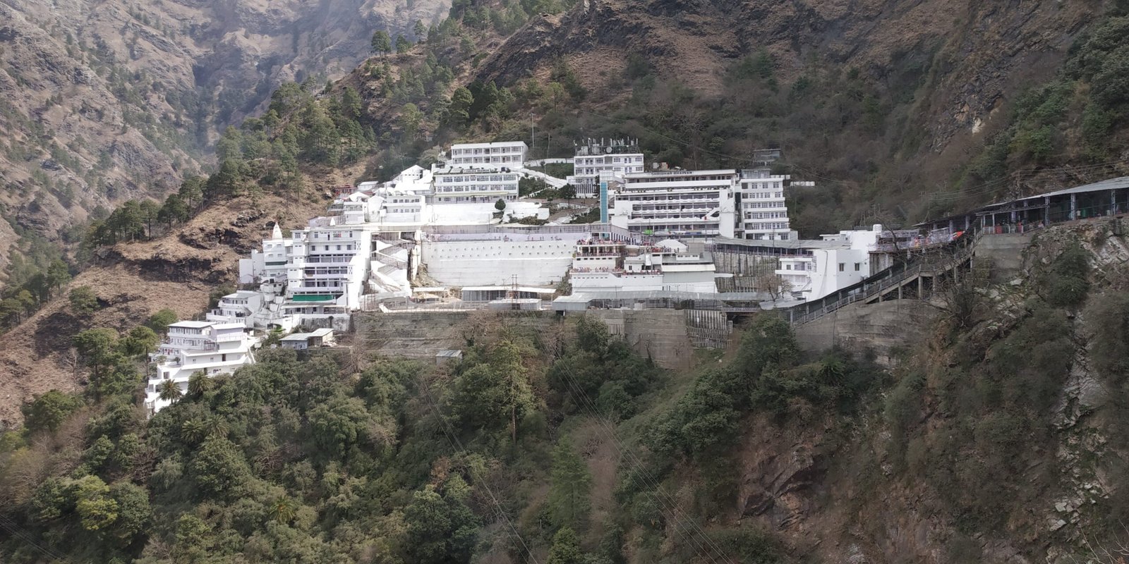 White buildings of Vaishno Devi Bhawan extending along a green mountainside above a steep valley.