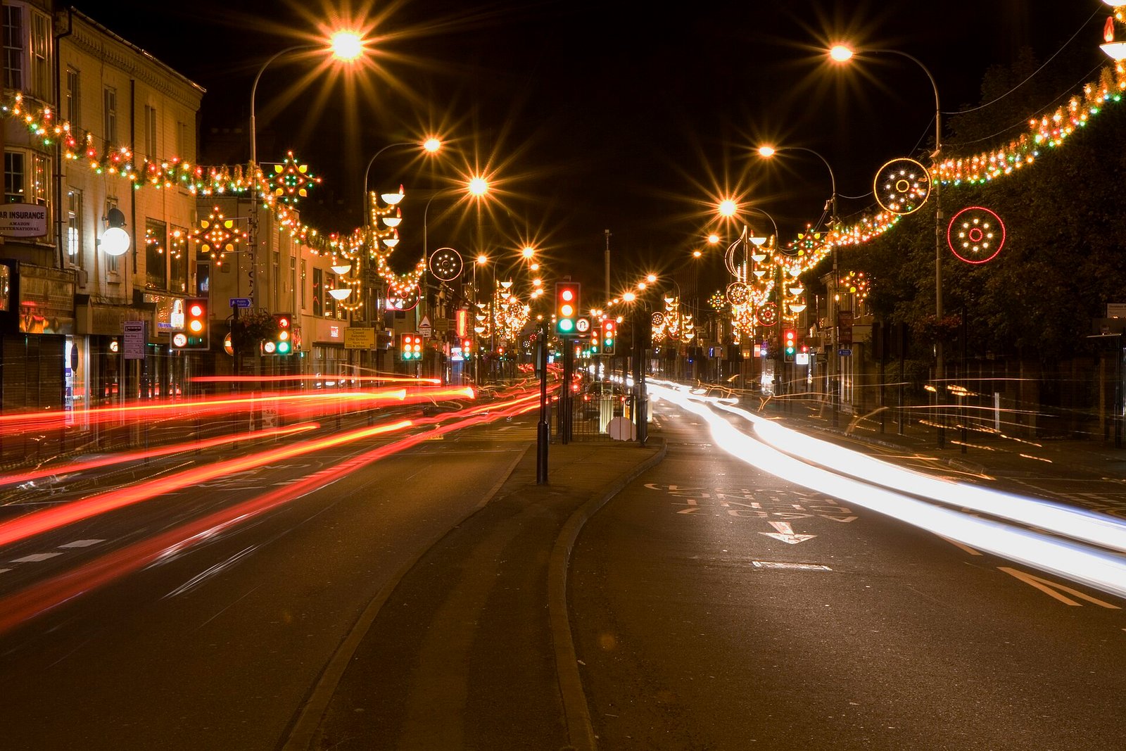 Lamp-shaped festive lights hang over Belgrave Road in Leicester, above shops and long streaks of passing traffic.