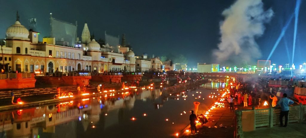 Visitors stand and tend rows of lamps along an Ayodhya waterfront, with illuminated buildings and reflections across the water.