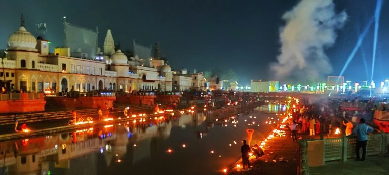 Visitors stand and tend rows of lamps along an Ayodhya waterfront, with illuminated buildings and reflections across the water.