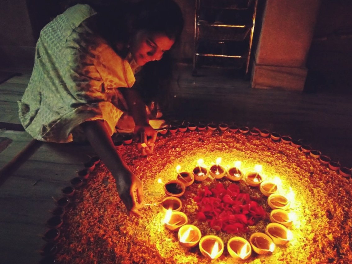 A person bends to light earthen lamps arranged in rings around a flower-petal rangoli at a doorway.