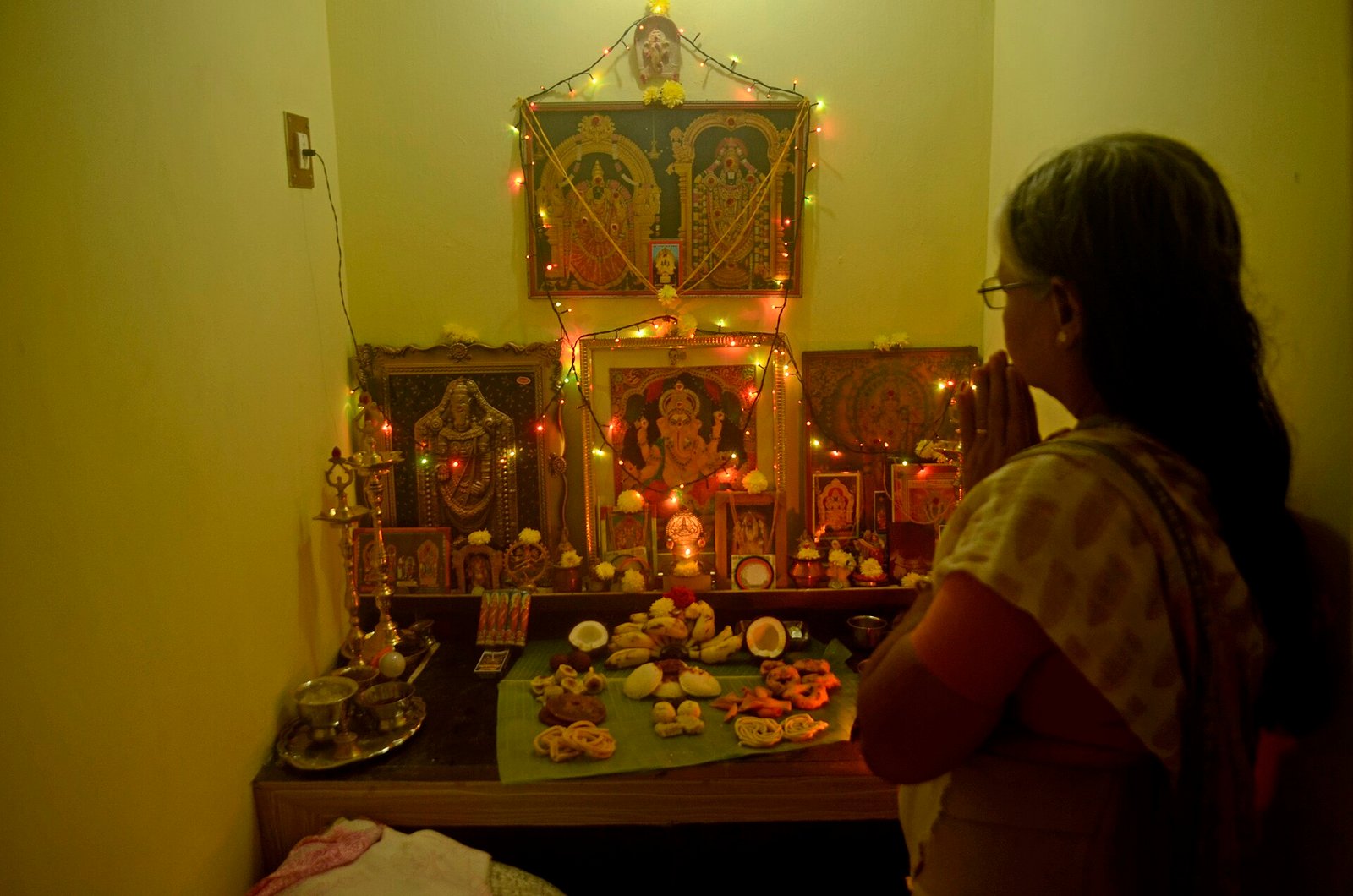 A worshipper joins her palms before a household shrine decorated with lights, with sweets, bananas and coconut arranged on banana leaves.