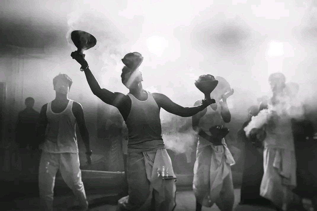 A participant holds smoking dhunuchi incense burners in both hands while others stand behind him in a smoke-filled festival setting.