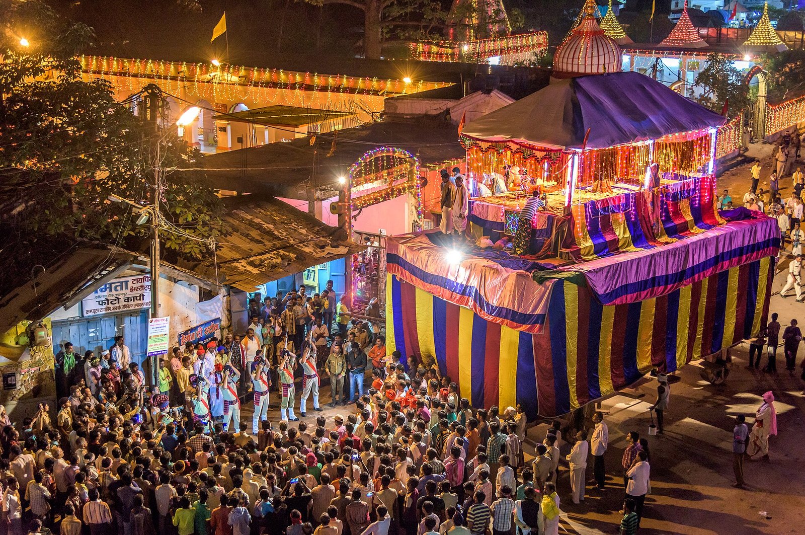People and musicians gather around a tall, illuminated chariot covered with purple, yellow and red fabric on a street at night.