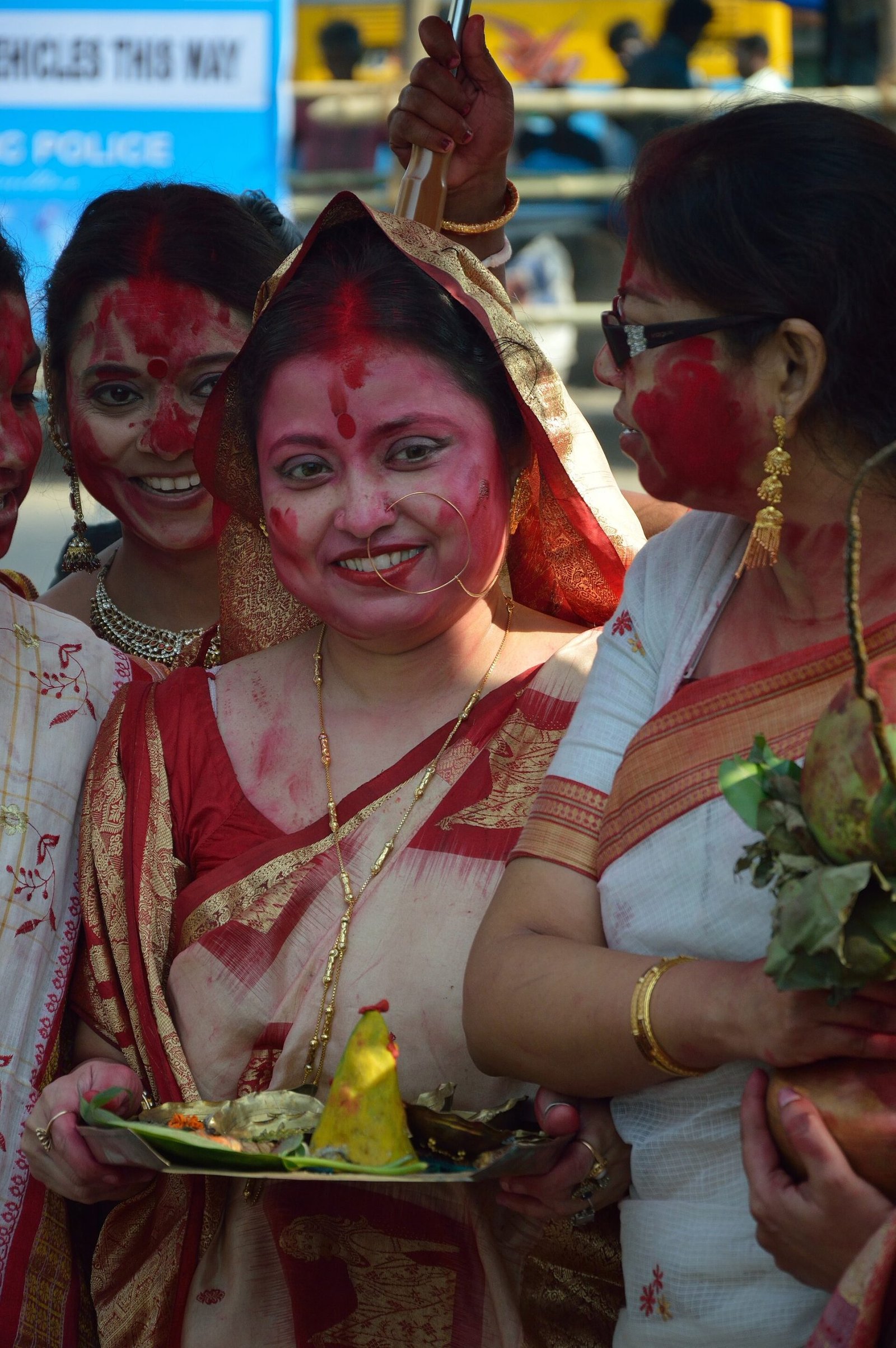 Women in white-and-red saris, with vermilion on their faces and hair partings, gather beside ritual offerings.