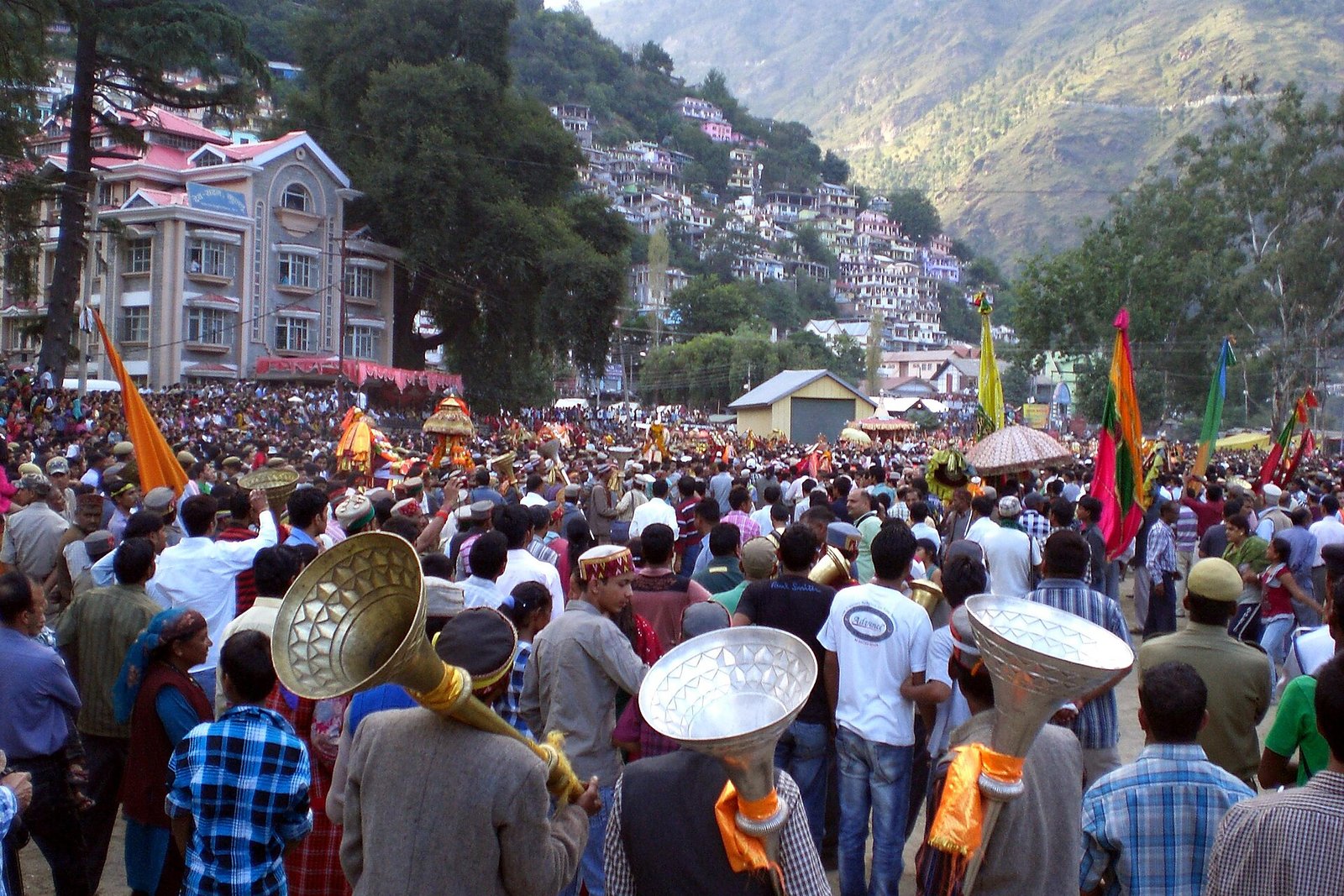 Musicians carry large flared brass instruments among a crowd and decorated palanquins, with Kullu hillside buildings behind them.