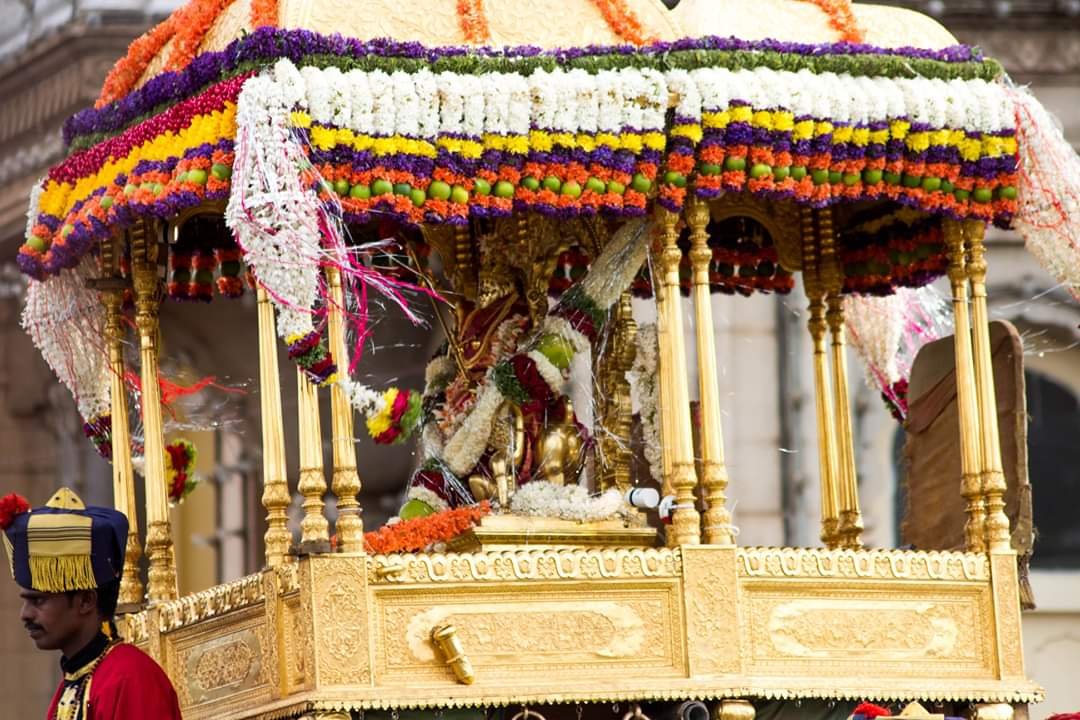 A flower-covered Goddess image sits inside a golden howdah beneath a striped canopy of flower garlands, with attendants alongside.