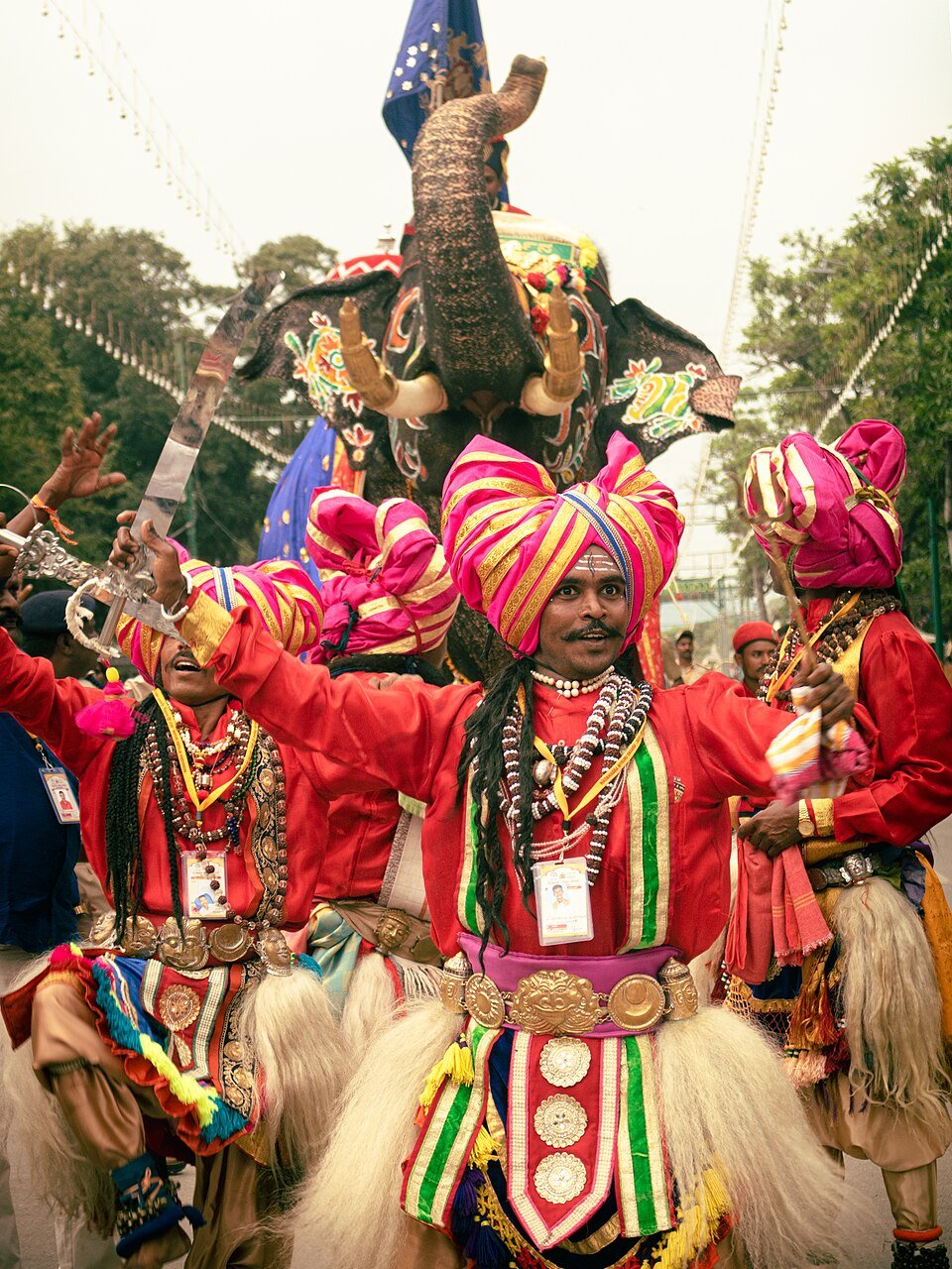 Performers in red clothing and pink-and-yellow turbans raise their arms, with a decorated elephant behind them.