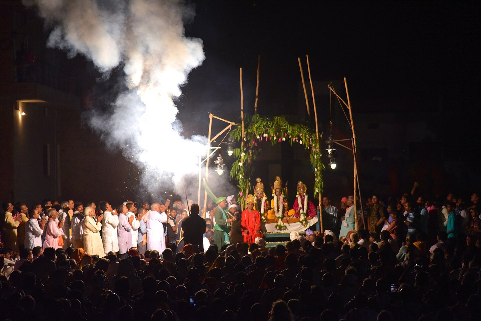 A nighttime audience surrounds crowned Ramlila performers beneath a leafy canopy, beside rising illuminated smoke.