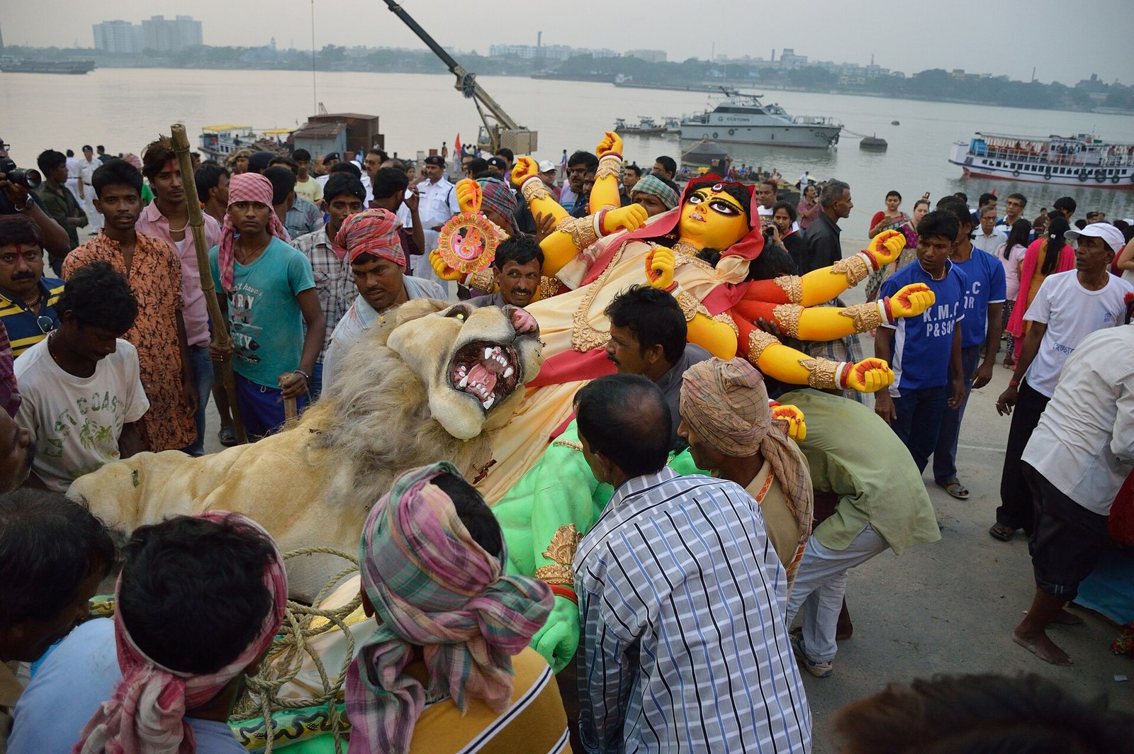 Participants carry a many-armed Durga image and lion toward the Hooghly at Baja Kadamtala Ghat in Kolkata, with boats on the river behind them.