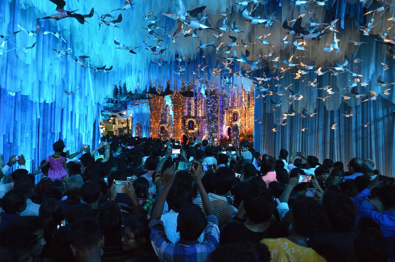 Visitors look into the Tridhara Sammilani Durga Puja pandal in Kolkata beneath blue-lit hanging fabric and suspended bird-shaped decorations.