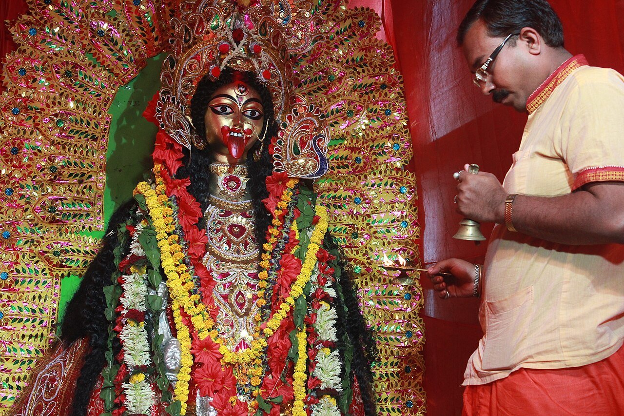 A man holding a bell and incense beside a flower-adorned Kali image during Kali Puja in Kolkata.