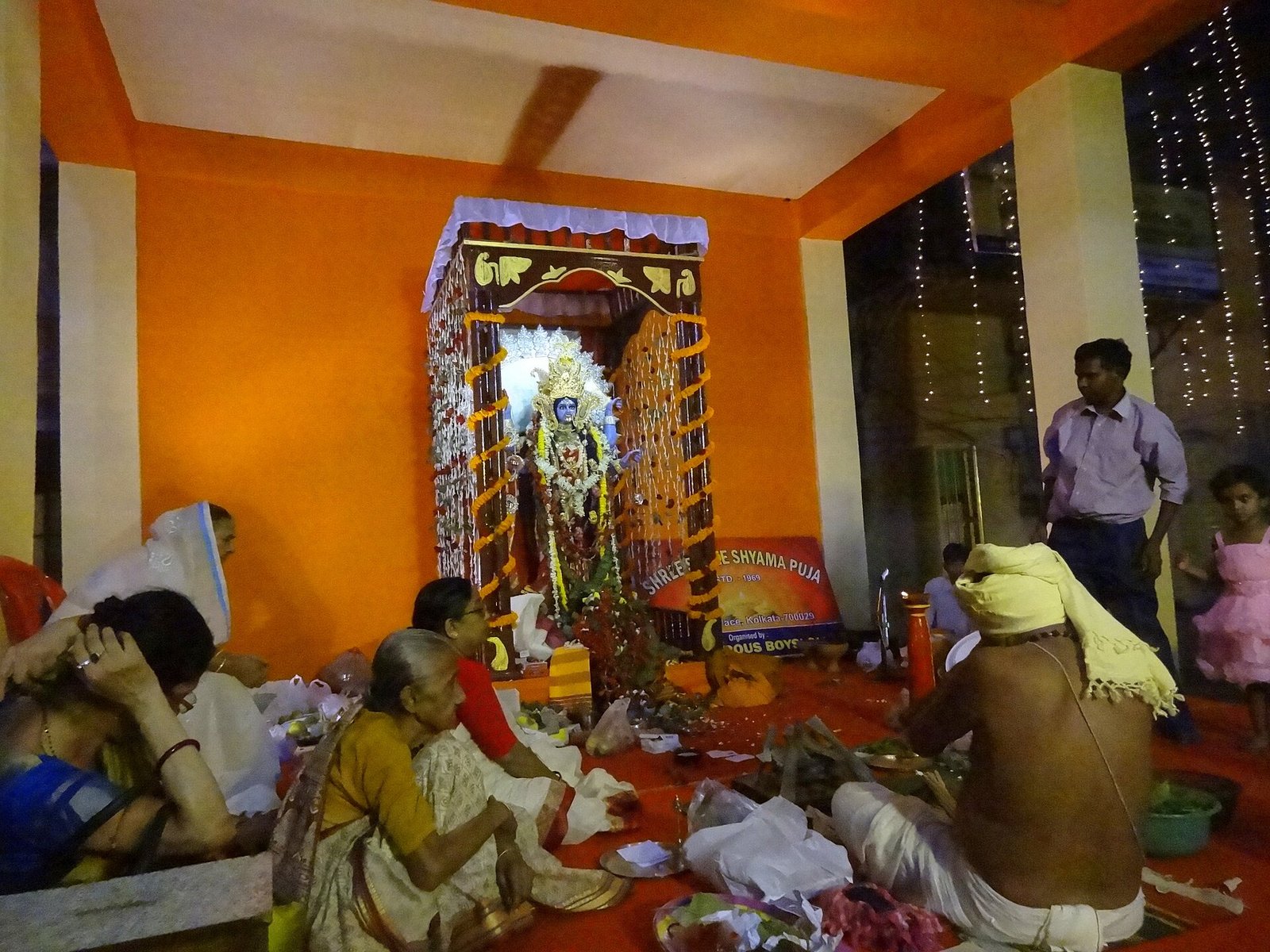 Worshippers sit beside offerings as a priest faces a garlanded Kali image in an illuminated neighborhood shrine.
