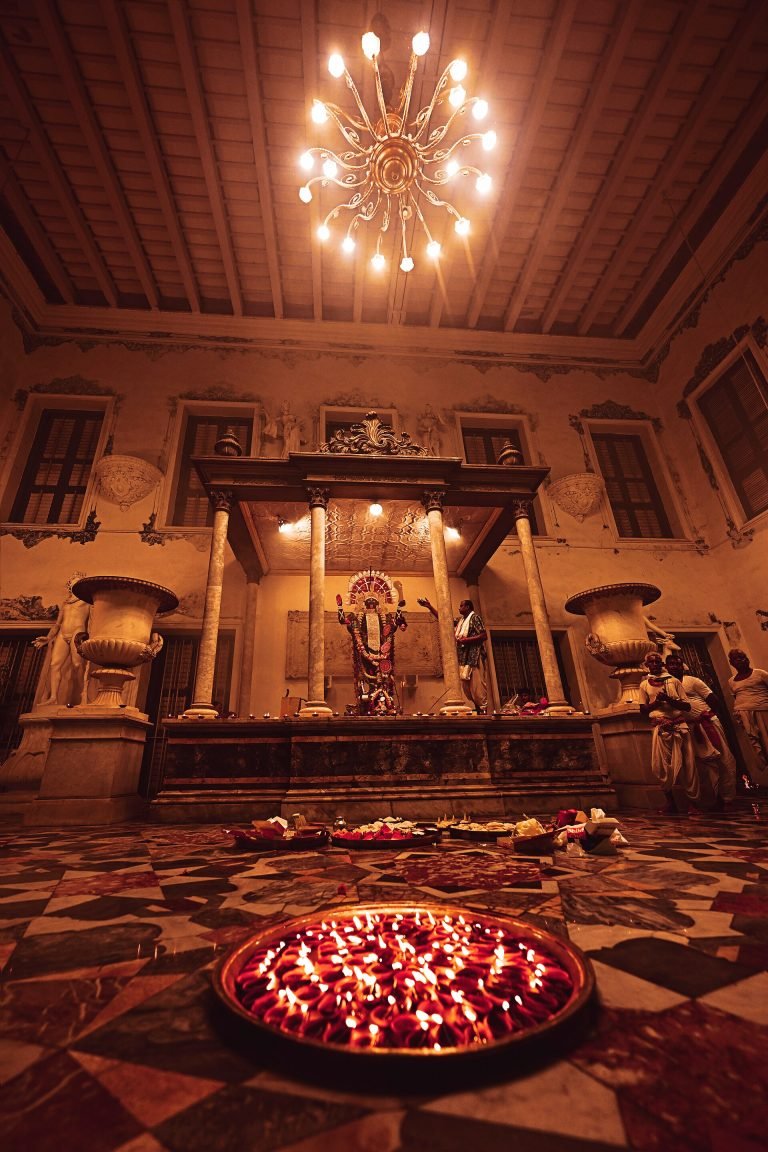 A tray of burning lamps leads toward a garlanded Kali image and worshippers beneath a chandelier in the Marble Palace courtyard.