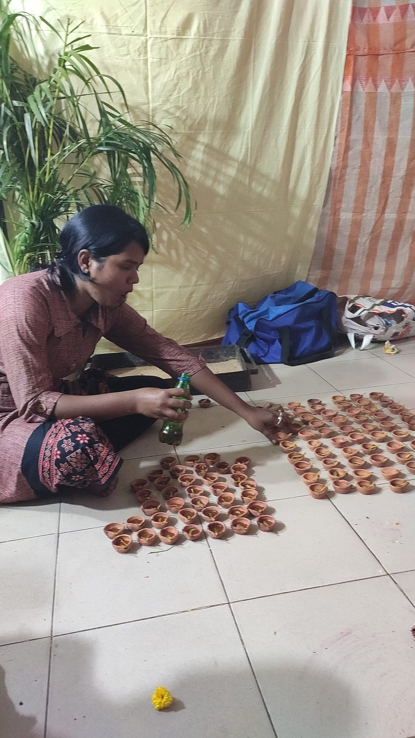 A seated woman prepares rows of small earthen lamps on the floor, holding a bottle beside the arranged wicks.