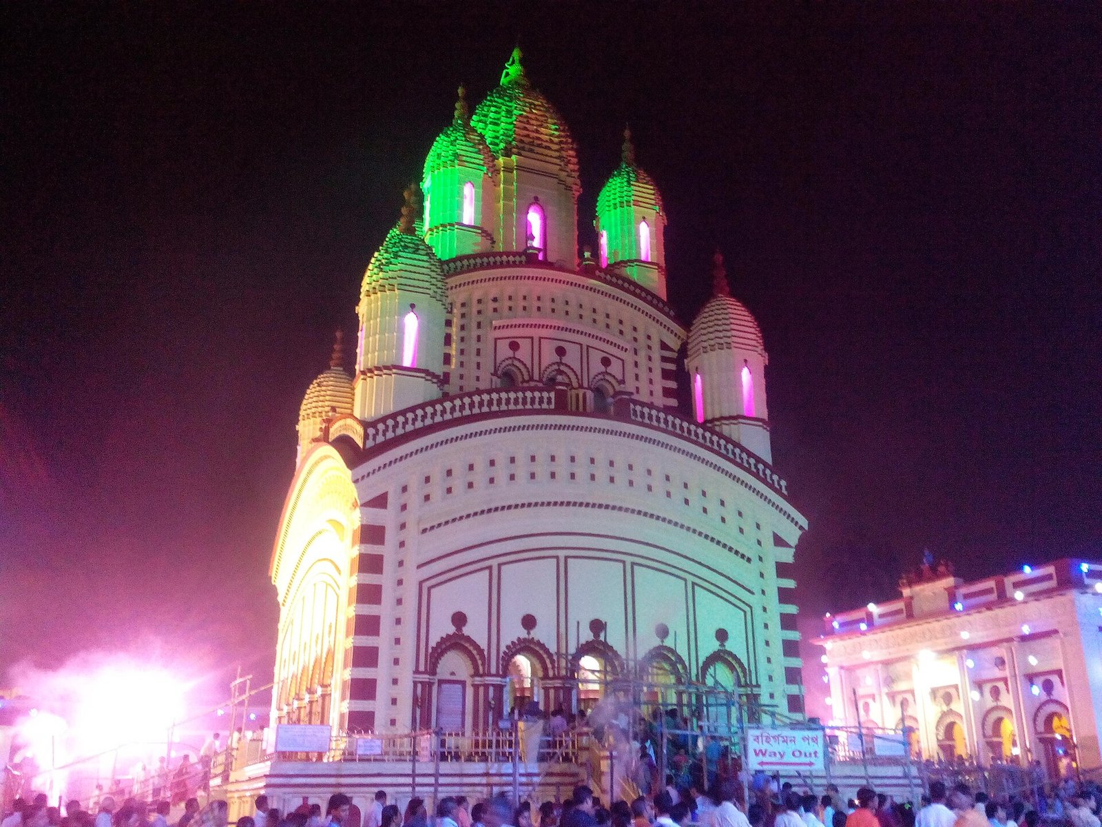 Visitors gather below the towers of Dakshineswar Kali Temple, illuminated in green and pink against the night sky.