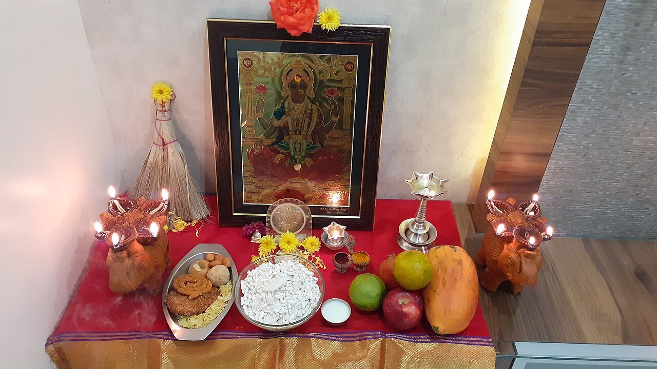Household Lakshmi Puja arrangement with a framed Lakshmi image, lit lamps, flowers, fruit and food offerings on a red cloth.