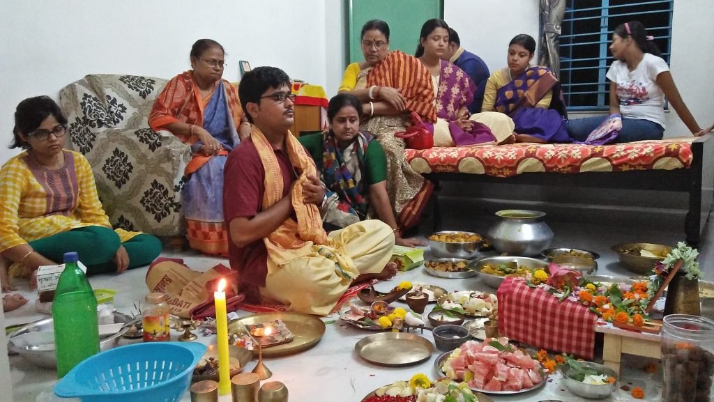 Family members sit around a man with joined palms, with a burning candle, flowers, vessels and food offerings arranged before a Lakshmi shrine.