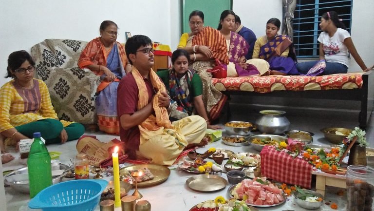 Family members sit around a man with joined palms, with a burning candle, flowers, vessels and food offerings arranged before a Lakshmi shrine.