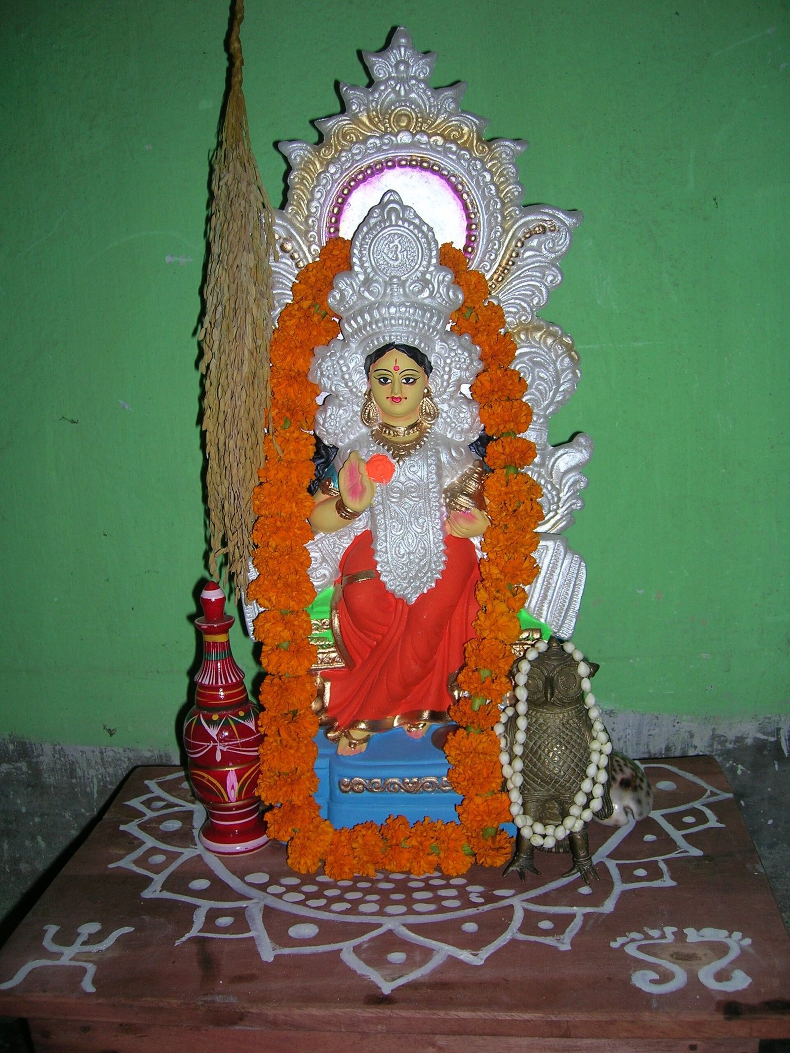 A Lakshmi image with marigold garlands stands beside grain stalks and ritual vessels on a platform decorated with white alpana patterns.