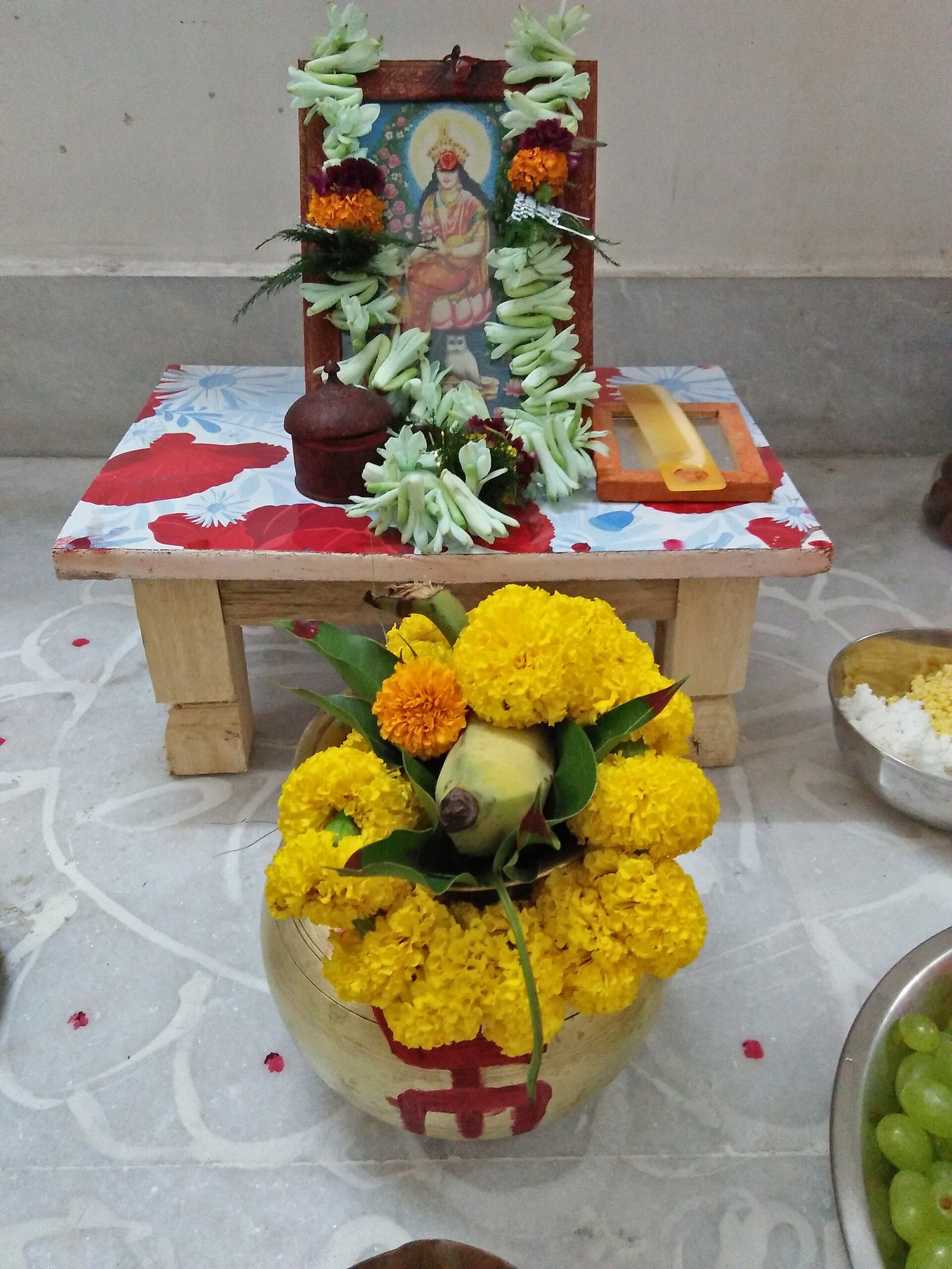 A garlanded Lakshmi picture stands above a flower-covered ritual vessel topped with a coconut, on a floor decorated with white alpana.
