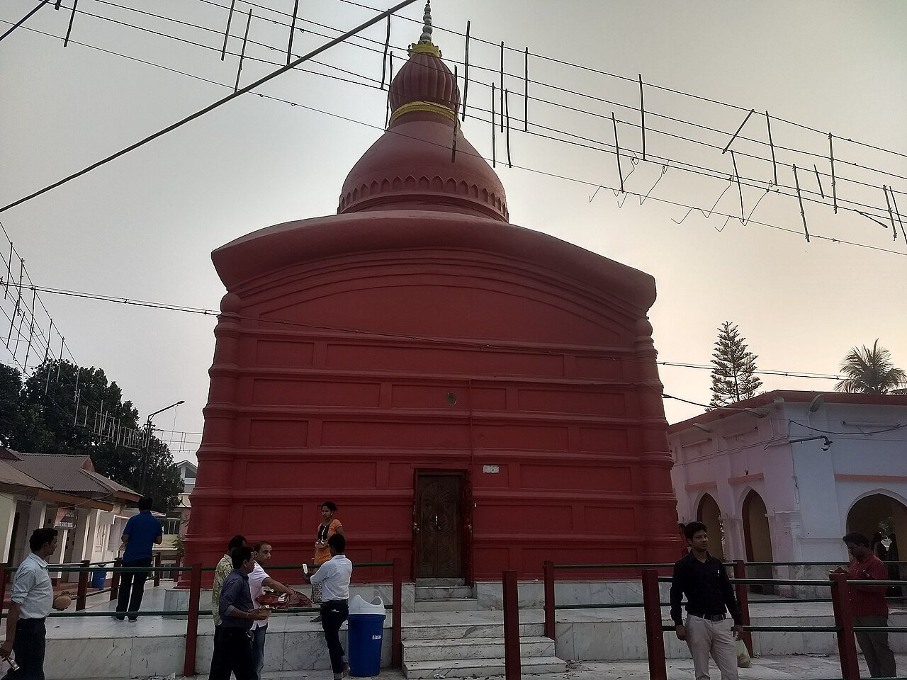 Red Tripura Sundari Temple at Matabari with its curved roof and rounded tower, visitors and courtyard railings in the foreground.