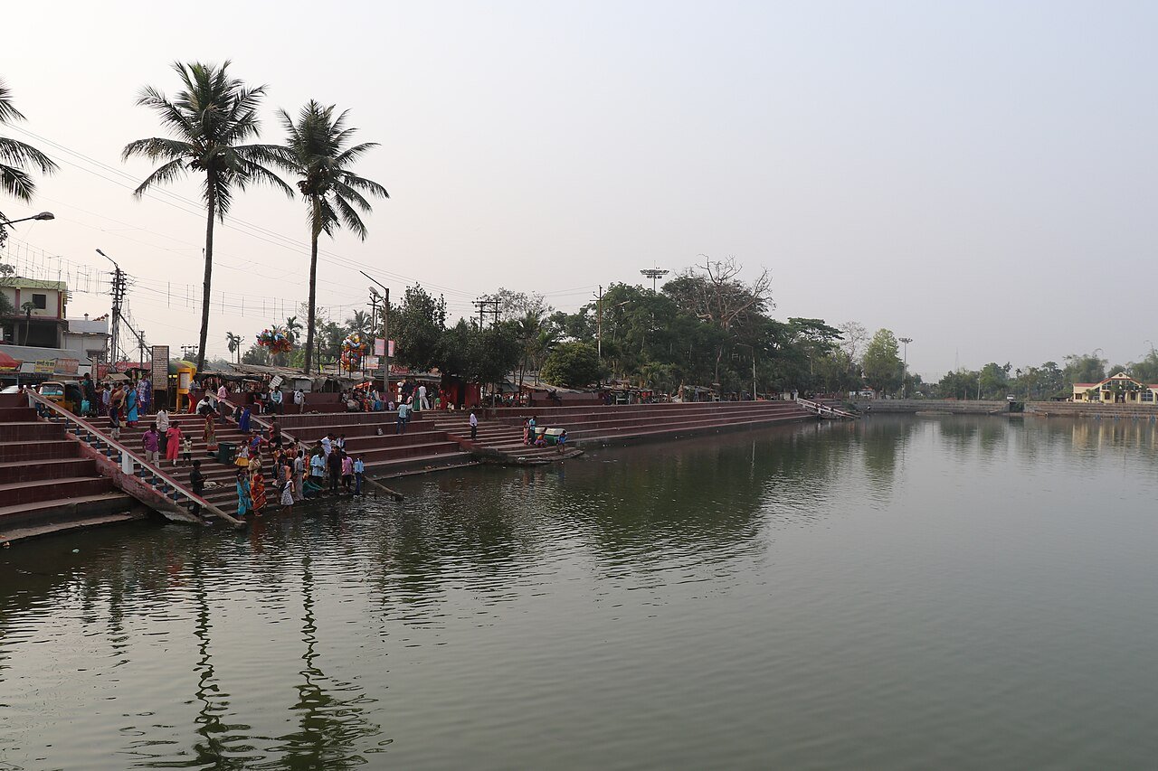Visitors gather on broad steps beside Kalyan Sagar at Matabari, with palms and stalls along the bank and the pond stretching into the distance.
