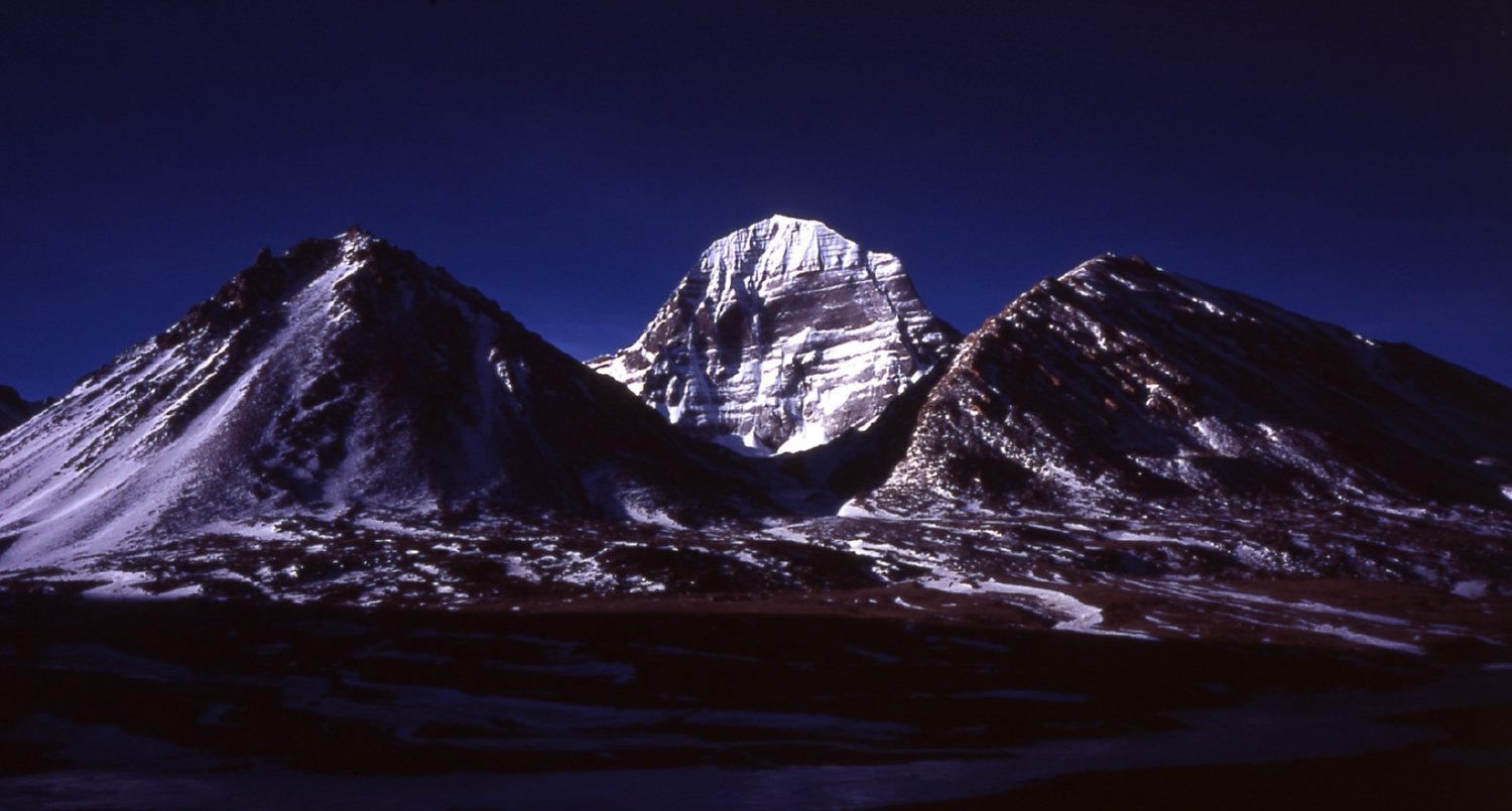 Snow-covered north face of Mount Kailash in Tibet beneath a deep blue sky, with dark ridges in the foreground.