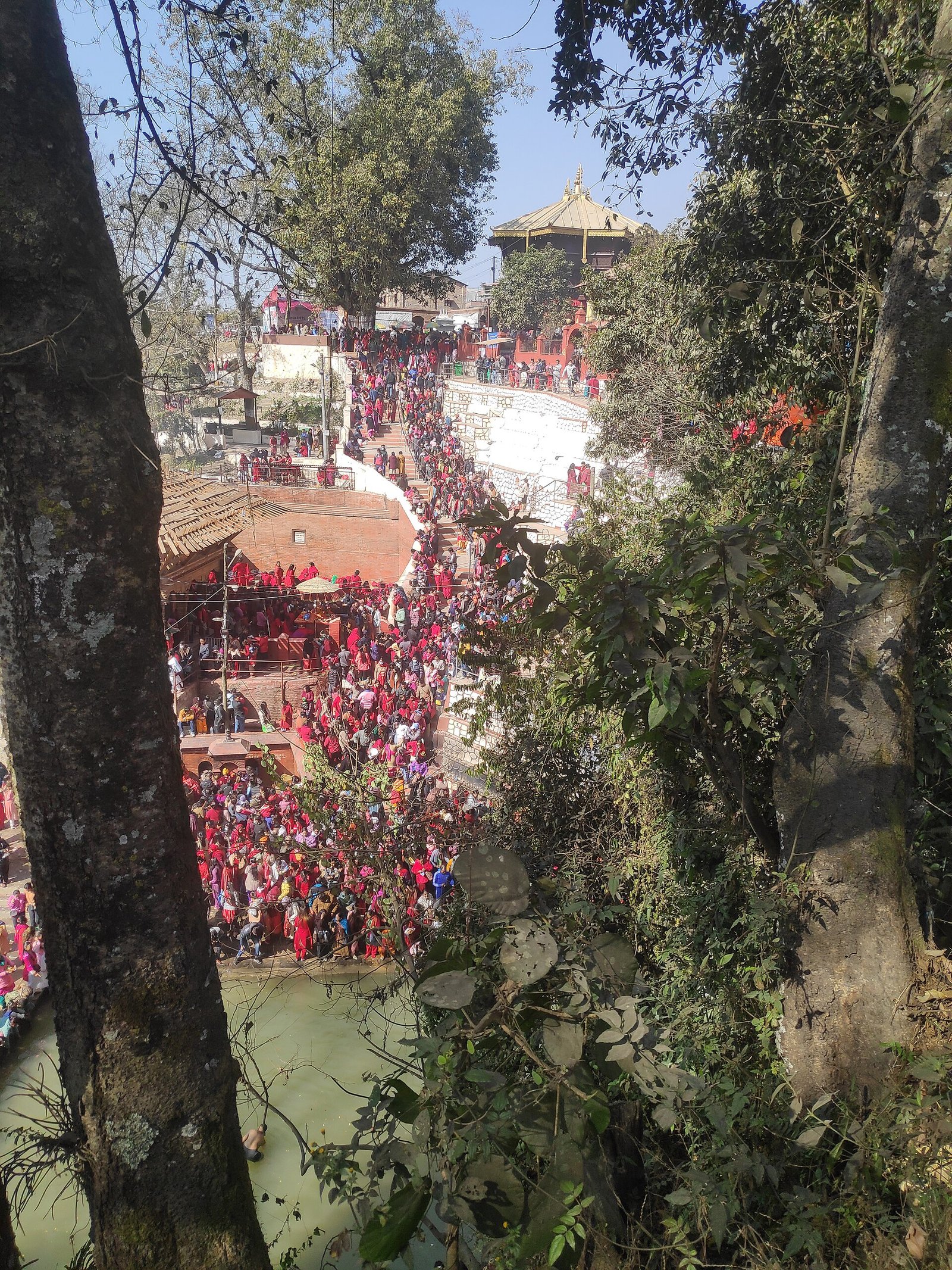 Crowds in red and other colorful clothing gather beside the river and on temple steps during Sali Nadi Mela at Sankhu, Nepal.