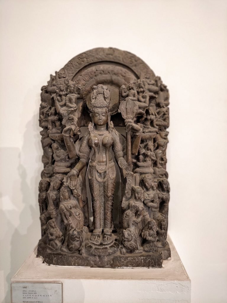 Standing stone Parvati wearing a tall crown and jewelry, framed by an ornate carved arch and smaller figures, at the National Museum in New Delhi.