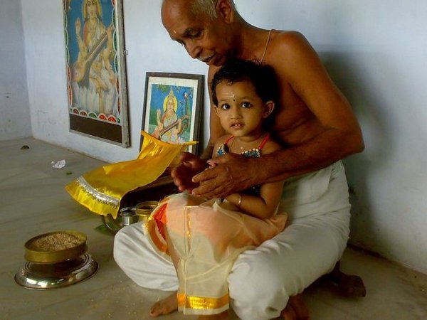 An adult holds a young child during Vidyarambham, beside a bowl of grain and framed Saraswati images.