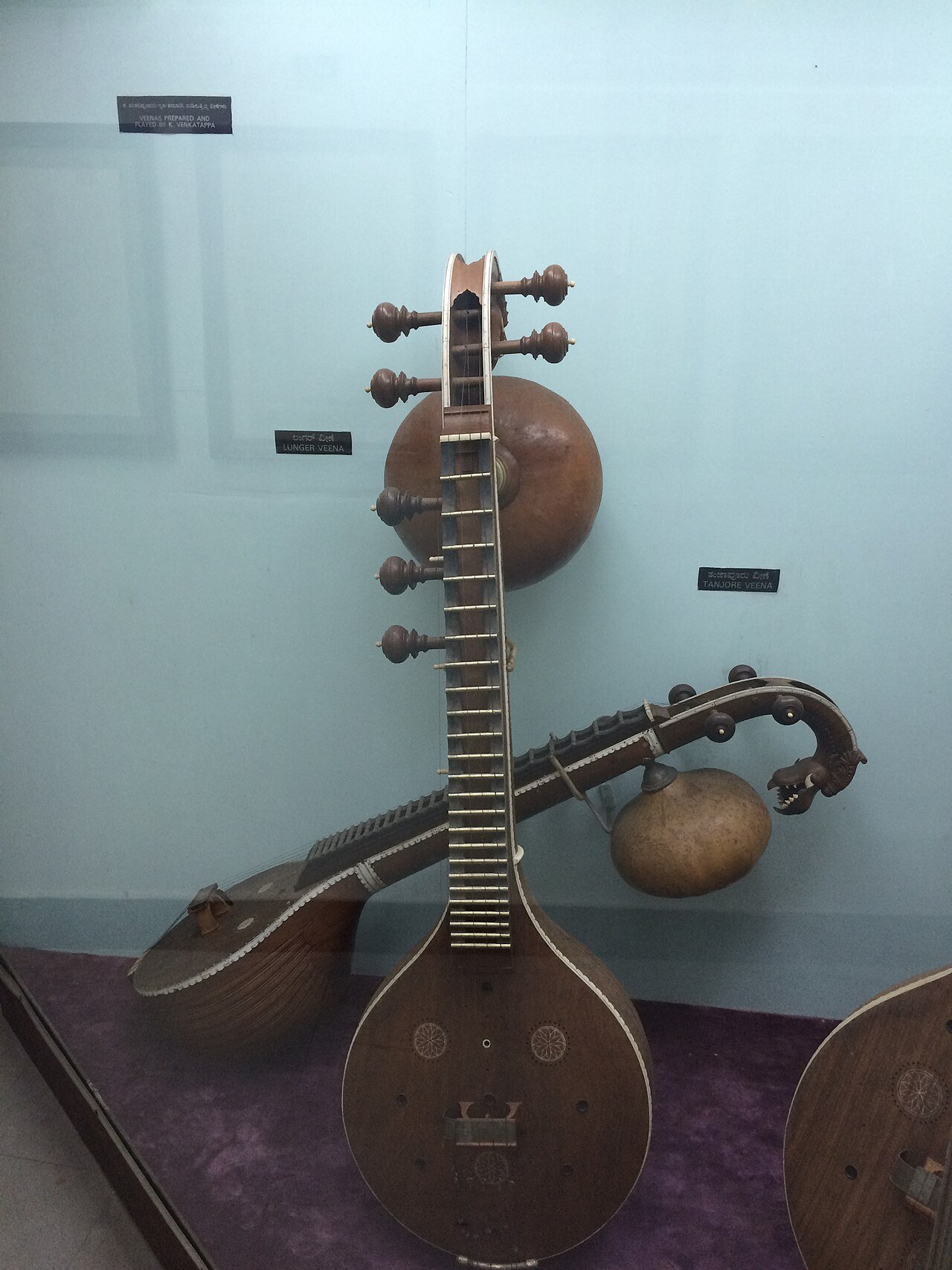Wooden veenas displayed in a museum case, showing long fretted necks, tuning pegs, rounded resonators and a carved animal-head terminal.