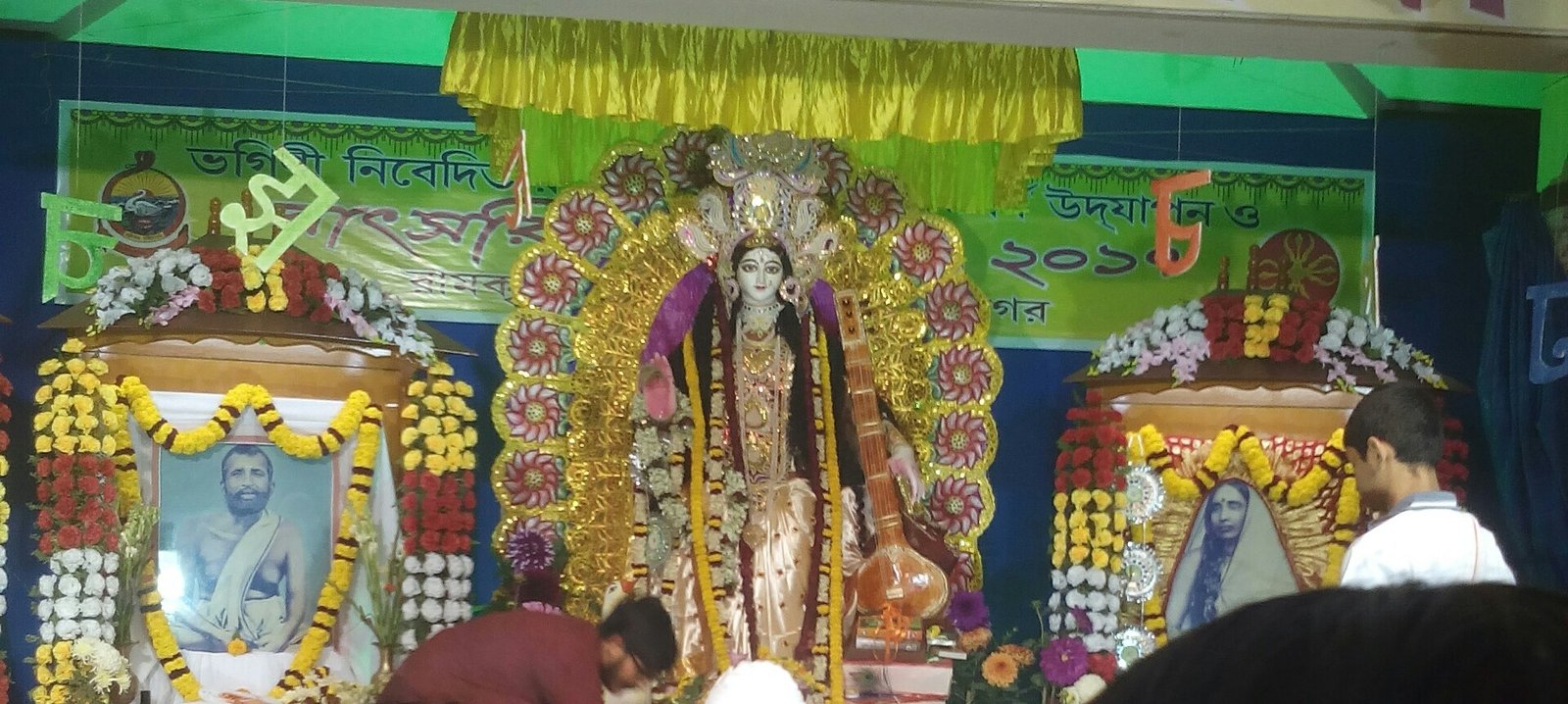 Flower-decorated Saraswati with a veena at a Kolkata school's puja, flanked by portraits of Ramakrishna and Sarada Devi, with attendees in front.
