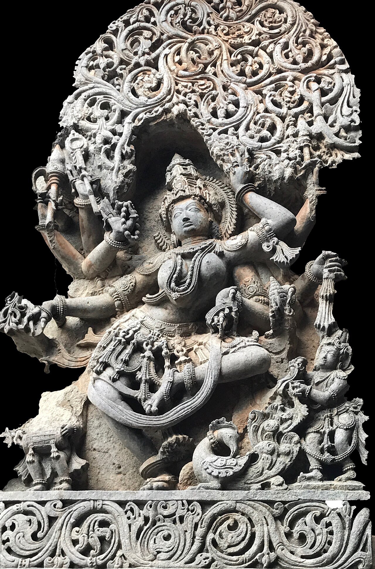 Carved stone Saraswati in a dancing pose beneath a foliage arch, with multiple arms holding attributes including a palm-leaf manuscript.